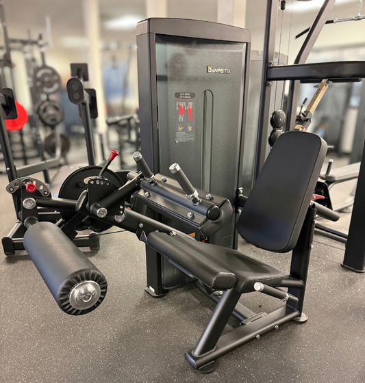 A Dynasty fitness leg extension leg curl machine in a gym setting, featuring a black adjustable bench, leg extension roller, and weight stack frame on a speckled gray floor.