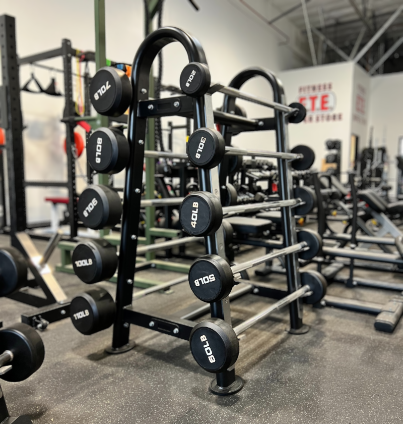 A black and silver fixed straight barbell with marked weight plates displayed on a black metal rack in a gym setting