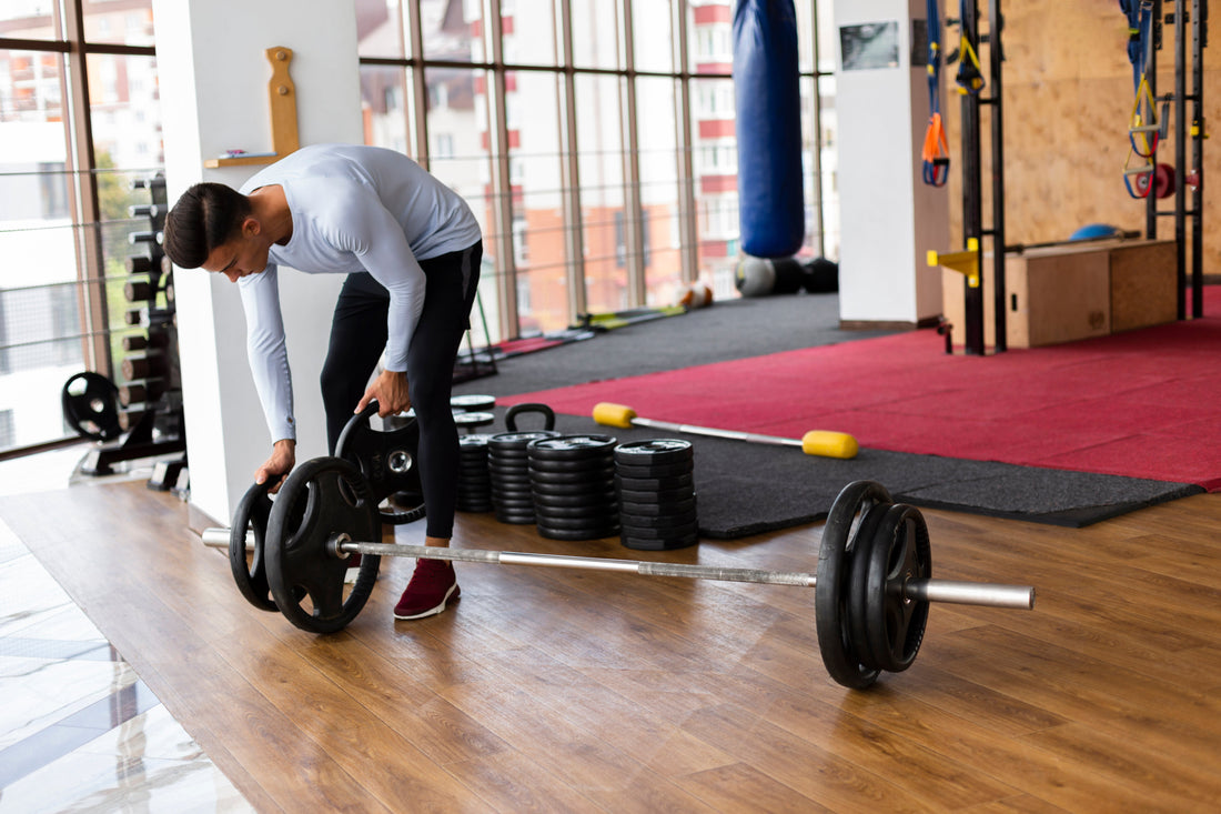 A man in athletic clothing places weight plates onto a barbell in a well-lit gym with wooden flooring, stacked plates, kettlebells, and training equipment in the background.