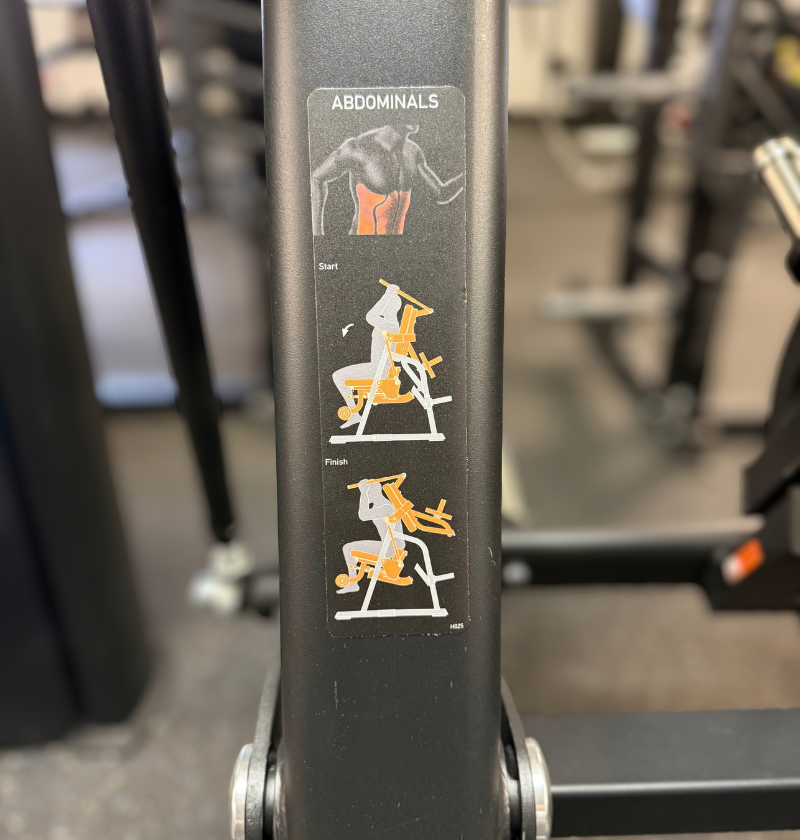 A close-up of a black ab crunch machine frame with a vertical instructional sticker labeled 'ABDOMINALS' showing exercise steps with white and orange illustrations.
