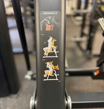 A close-up of a black ab crunch machine frame with a vertical instructional sticker labeled 'ABDOMINALS' showing exercise steps with white and orange illustrations.