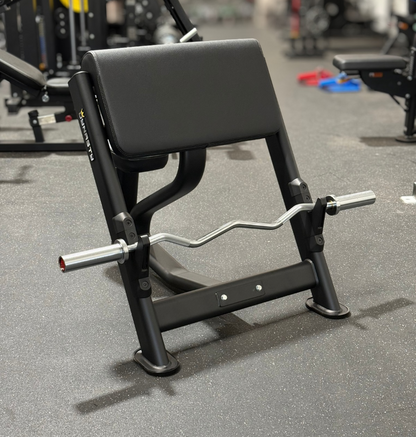 A black preacher bench with a curl barbell, photographed in a gym setting with other exercise equipment blurred in the background.