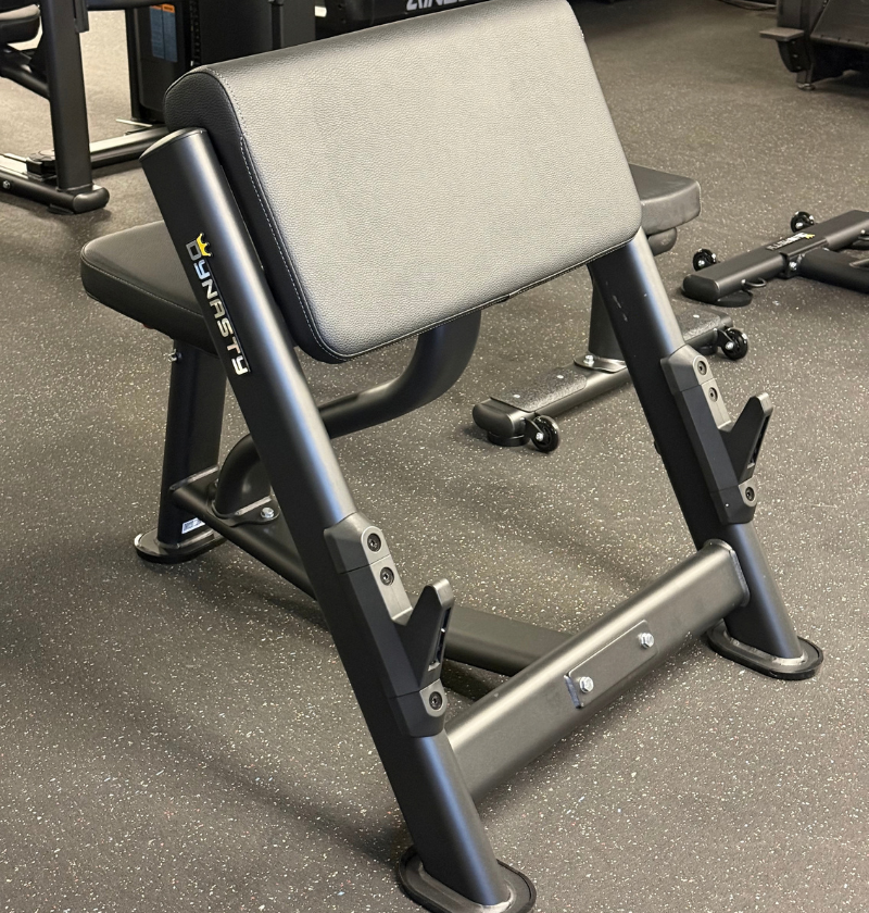 A preacher bench in black and gray, photographed in a gym setting on a speckled gray floor.