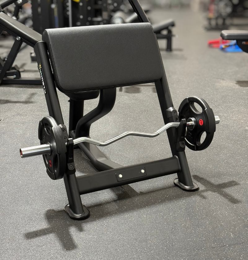 A black preacher bench with a curl barbell loaded with black weight plates, photographed in a gym setting on a gray floor.