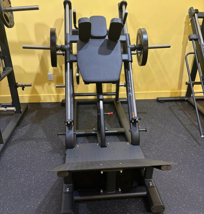A black hack squat gym machine with weight plates, set in a gym with a yellow wall and dark speckled floor.
