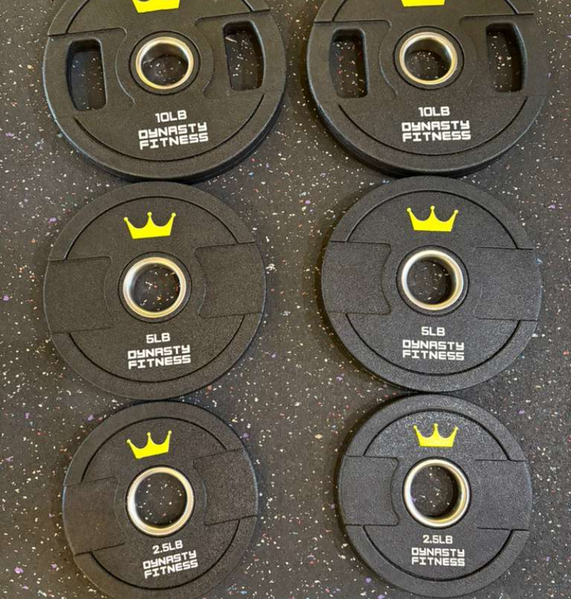 Set of black urethane weight plates with yellow crowns and 'Dynasty Fitness' branding on a dark speckled surface.